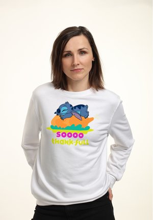 Kobieta o brązowych włosach do ramion, ubrana w białą bluzę z nadrukiem śpiącej niebieskiej postaci z kreskówki na pieczonym indyku z napisem "TAK BAAAAARDZO WDZIĘCZNA".