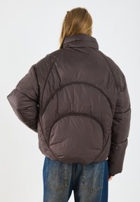 Persoană cu păr lung, purtând o geacă puf dintr-un material închis la culoare, cu cusături circulare pe spate, și blugi din denim albastru, întoarsă cu spatele.