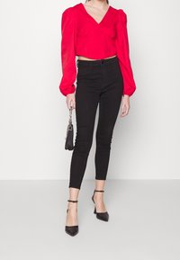 Blusa roja corta con mangas abullonadas, escote en forma de V, combinada con jeans ajustados negros y tacones negros de punta. Pequeño bolso negro con cadena.