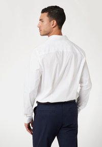 Camisa blanca de manga larga con detalle en el yugo trasero, corte ajustado, puños con botones y textura de tela suave. Llevada con pantalones azul marino.