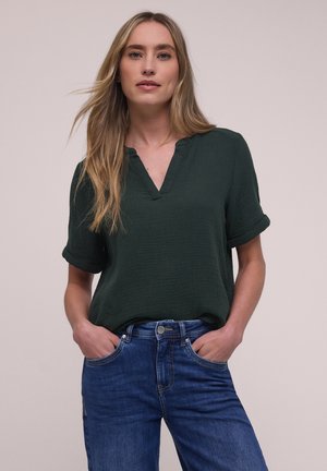 Frau mit langem blondem Haar, die eine dunkelgrüne kurzärmelige Bluse und blaue Jeans trägt, stehen mit den Händen in den Taschen vor einem schlichten Hintergrund.