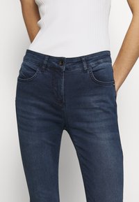 Jeans ajustados de denim azul oscuro, con cierre de botón en la parte delantera, trabillas para el cinturón y bolsillos laterales con sutil detalle de costura.