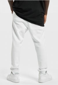 White sweatpants with a relaxed fit, elastic cuffs, and side pockets, paired with white sneakers, viewed from the back. Black shirt visible.