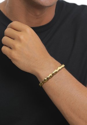 Mann mit schwarzem Hemd zeigt ein goldenes, gedrehtes Armband am linken Handgelenk, Hand nahe der Schulter vor weißem Hintergrund.