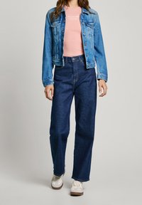 Denim jacket in light blue with two chest pockets, paired with a pink fitted t-shirt and high-waisted dark blue jeans. White sneakers.