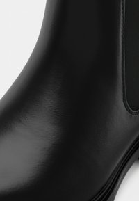 Μαύρη καουτσούκ μπότες έως τον αστράγαλο με γυαλιστερή επιφάνεια, ελαστικές πλαϊνές επιφάνειες και λεία υφή. Στρογγυλή μύτη και διακριτική λεπτομέρεια ραφής.