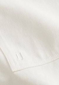 White textured fabric with a subtle ribbed pattern, featuring a small embroidered "U" logo in white. Clean lines and minimal detailing.
