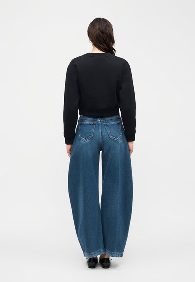 Γυναίκα που στέκεται με την πλάτη, φορώντας μαύρο μακρυμάνικο τοπ, φαρδιά μπλε τζιν παντελόνια και μαύρα παπούτσια, μπροστά σε απλό φόντο.