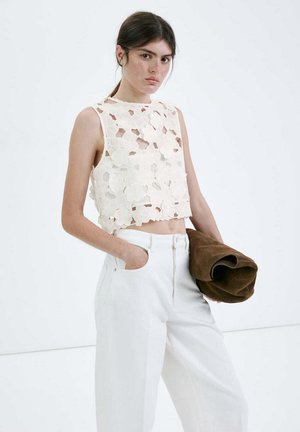 Giovane donna con capelli scuri indossa un crop top bianco in pizzo floreale senza maniche e pantaloni bianchi a vita alta, tenendo una giacca marrone piegata.