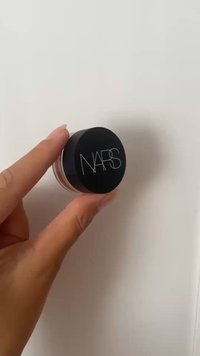Produit crémeux NARS dans un petit pot transparent avec un couvercle noir mat portant des lettres blanches. Forme ronde et slim, tenu entre les doigts.