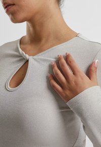 Lys grå langermet topp med et strukturert, glitrende stoff. Har en nøkkelhullutskjæring og en stilig knebånd-detalj ved halsen.