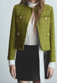 Chaqueta de tweed texturizada verde con botones dorados, dos bolsillos frontales y un diseño corto, combinada con una blusa blanca y una falda negra.