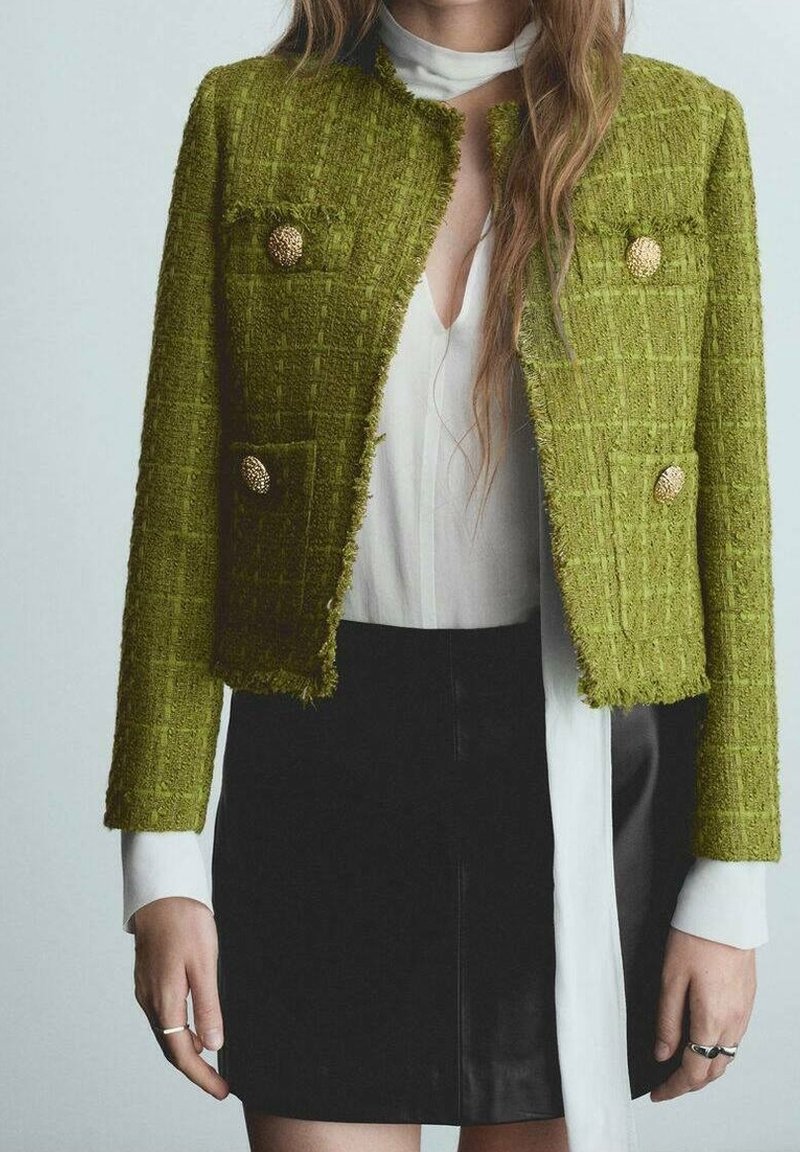 Chaqueta de tweed texturizada verde con botones dorados, dos bolsillos frontales y un diseño corto, combinada con una blusa blanca y una falda negra.
