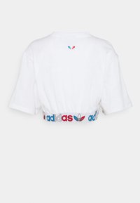 Bílý zkrácený tričko s krátkým rukávem a žebrovaným pasem. Na zadní straně jsou multibarevné logo "adidas" a ikonický design se třemi pruhy.