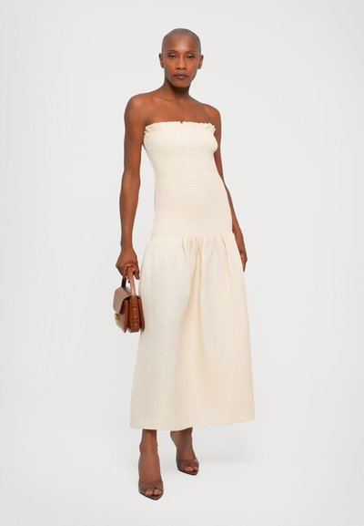 Trägerloses, beiges Kleid mit einem gerafften Oberteil und fließendem, plissiertem Rock. Das Model hält eine braune Handtasche und trägt Sandalen mit offenem Zehenbereich.