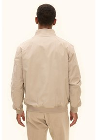 Beige Bomberjacke mit Stehkragen, gerippten Bündchen und glatter Textur. Verfügt über Nahtdetails und eine lockere Passform. Rückansicht gezeigt.