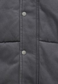 Materialul de puffer gri are un model matlasat, dotat cu nasturi cu presiune argintii pe față. Textură netedă și construcție robustă.