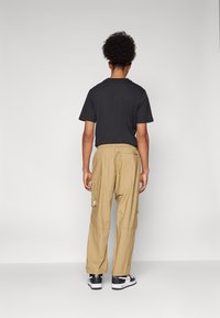 Fekete, rövid ujjú póló, bézs színű, elasztikus derékú cargo nadrág oldalzsebekkel, fekete-fehér sportcipőkkel viselve, hátulról nézve.