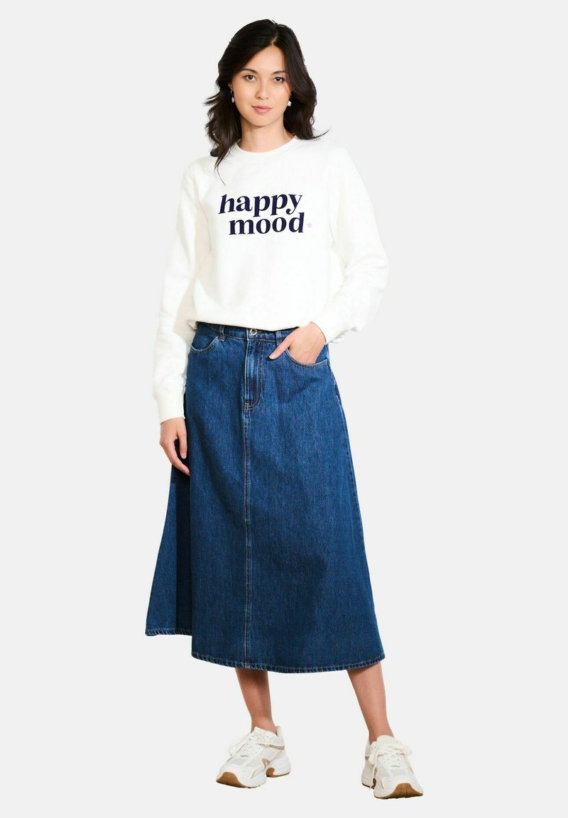 Witte sweatshirt met "happy mood" in marineblauw gedrukt, gecombineerd met een donkerdenim rok. Model draagt witte sneakers en heeft haar handen in de zakken van de rok.