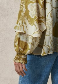 Bluză cu model floral în verde măsliniu și crem, cu mâneci bufante și manșete elastice. Asortată cu blugi de denim bleumarin.