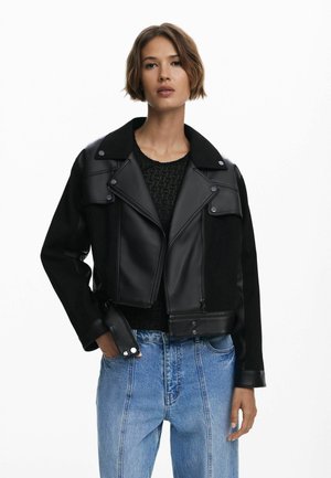 Black cropped jacket featuring leather and fabric contrast, pointed collar, front zip, and snap buttons. Paired with light blue denim jeans.