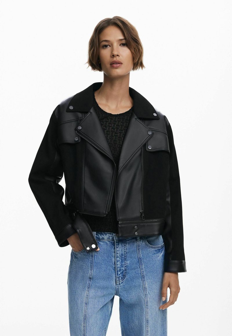 Zwarte cropped jas met een contrasterende combinatie van leer en stof, een puntige kraag, een rits aan de voorkant en drukknoopsluitingen. Gecombineerd met lichtblauwe denim jeans.