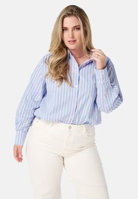 Chemise boutonnée bleu clair à rayures verticales roses et blanches, avec un col classique et des manches longues. Portée avec un pantalon blanc.
