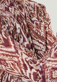 Blouse à manches longues, texturée, avec un col froncé. Présente un motif abstrait coloré en bordeaux, rose et vert sur un fond crème.