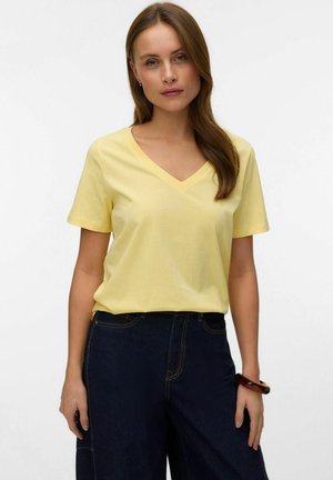 Woman with long brown hair wearing a light yellow V-neck T-shirt and dark blue jeans against a plain white background.