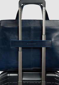 Maletín de cuero azul marino asegurado en el asa extensible de una maleta negra con ruedas, destacando la comodidad para viajar y los negocios.