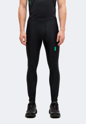 Άνδρας με μαύρο εφαρμοστό αθλητικό κολάν και μαύρα παπούτσια τρεξίματος στέκεται μπροστά σε φωτεινό φόντο.