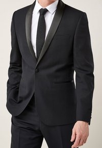 Veste de smoking noire avec revers en satin, fermeture à un bouton, et deux poches avant, portée sur une chemise blanche et une cravate texturée.