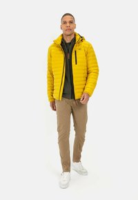 Gelbe Pufferjacke mit horizontalen Nähten, schwarzen Reißverschlüssen und Kapuze. Trägt sie über einem grünen Oberteil, kombiniert mit beigen Hosen und weißen Sneakers.