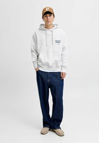 Sudadera gris claro con un bolsillo frontal y logo, combinada con unos vaqueros holgados de color azul oscuro. Zapatillas de tono neutro sin cordones. Gorra marrón.