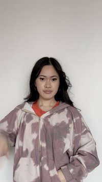 Ung kvinde med langt mørkt hår iført øreringe i form af ringe og en lilla og hvid tie-dye lynlåshættejakke over en orange top mod en ensfarvet baggrund.
