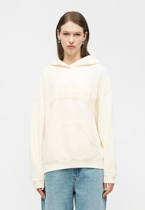 Νεαρή γυναίκα που φοράει κρεμ φούτερ με κείμενο "Blanche Blanche" με υφή, ασημένια σκουλαρίκια και ανοιχτό μπλε τζιν, στέκεται μπροστά σε λευκό φόντο.