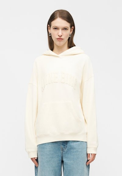 Jeune femme portant un sweat à capuche crème avec le texte texturé "Blanche Blanche", des boucles d'oreilles argentées et un jean bleu clair, debout devant un fond blanc.