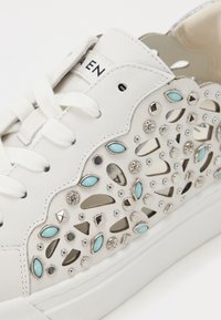 Hvit lær sneakers med utskjæringsdesign og turkise detaljer, metalliske stifter og flate snørebånd. Teksturer overflate med dekorative elementer.