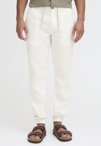 Pantaloni beige leggeri a taper con polsini elastici e vita con cordoncino. Abbinati a sandali marrone scuro. Texture liscia, design casual.