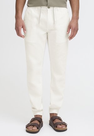 Pantaloni beige leggeri a taper con polsini elastici e vita con cordoncino. Abbinati a sandali marrone scuro. Texture liscia, design casual.