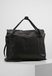 Schwarze Lederhandtasche mit silbernem Reißverschluss, doppelten Griffen und abnehmbarem Schultergurt, auf einem weißen Podest vor grauem Hintergrund platziert.