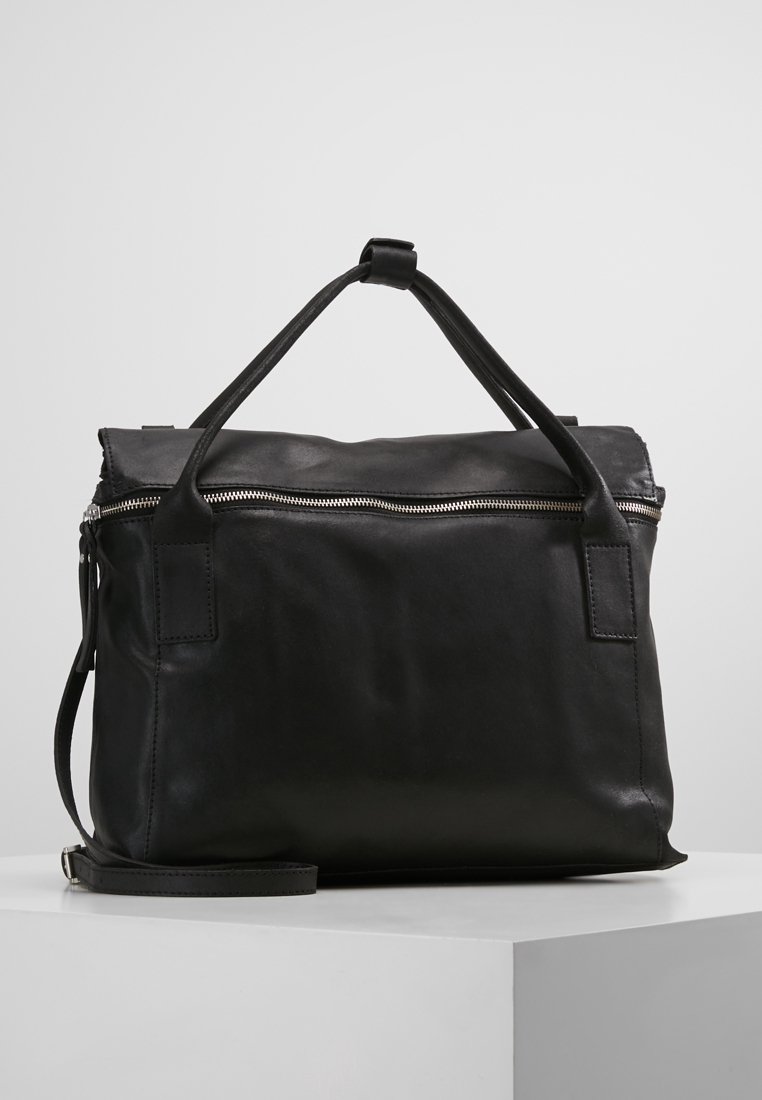 Schwarze Lederhandtasche mit silbernem Reißverschluss, doppelten Griffen und abnehmbarem Schultergurt, auf einem weißen Podest vor grauem Hintergrund platziert.