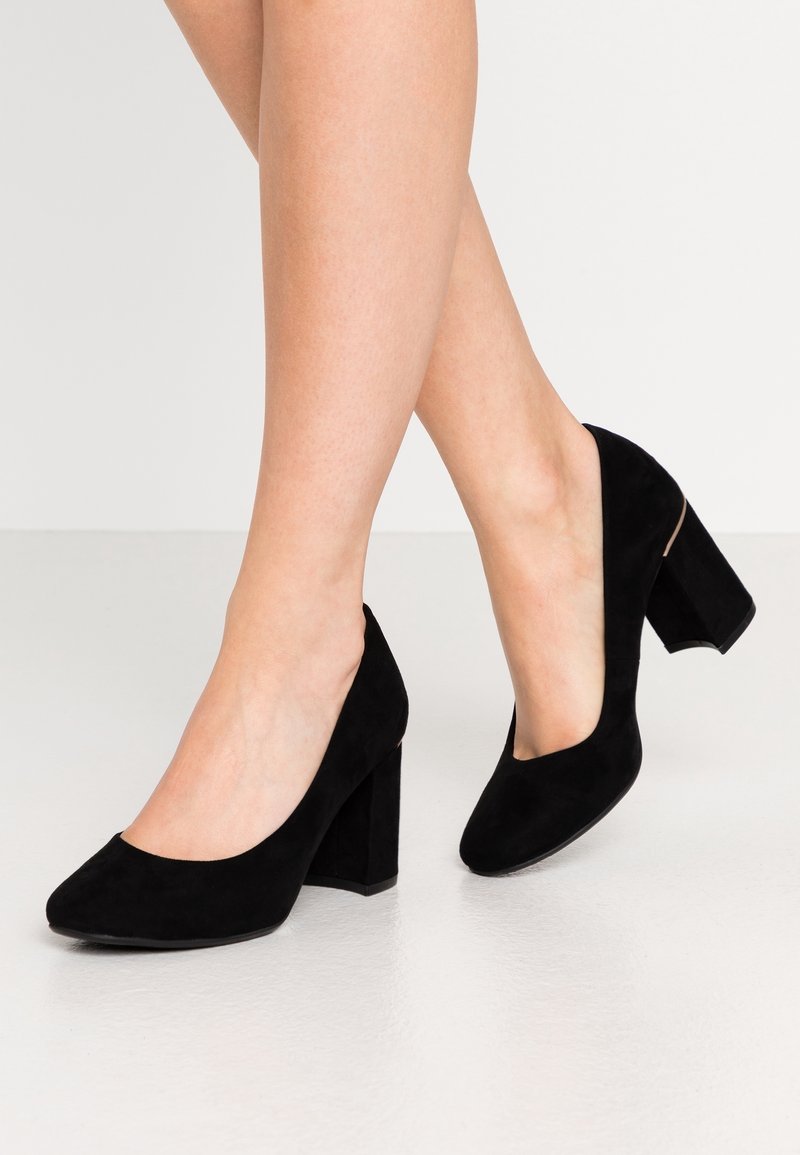 Jambes portant des chaussures en daim noir à talons carrés et bouts fermés sur un sol gris clair, fond blanc.