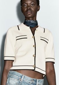 Top a maniche corte in maglia color crema con rifiniture nere e bottoni dorati. Presenta un colletto e due tasche frontali. Indossato con jeans in denim consumati.