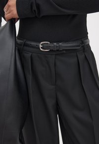 Czarny skórzany pasek z metalową, srebrną klamrą, noszony na czarnych spodniach z plisami i prążkowanej czarnej bluzce, prezentujący monochromatyczny design.