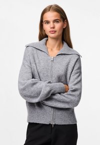 Young woman with long hair wearing a light gray zip-up sweater with wide collar, standing with arms crossed against a plain background.