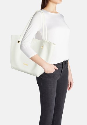 Donna con camicia bianca a maniche lunghe e jeans neri, con una grande borsa tote bianca strutturata con dettagli dorati sulla spalla.