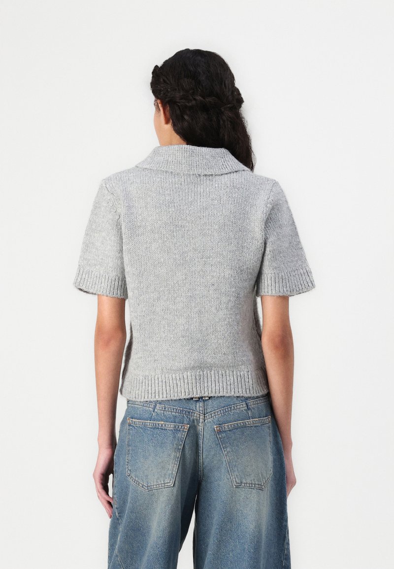 Pull en tricot gris à manches courtes avec un col et des poignets côtelés. Ourlet côtelé à la taille. Porté avec un jean léger bleu clair ample.