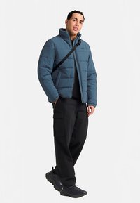 Giubbotto corto imbottito blu, con collo alto e chiusura con zip. Indossato sopra una camicia nera, abbinato a pantaloni cargo neri e sneakers.