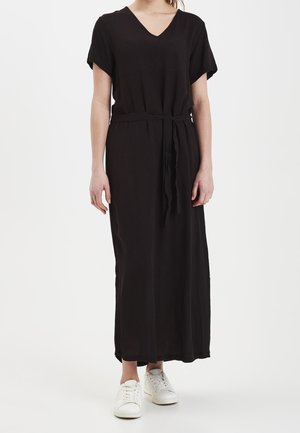 Woman wearing a black short-sleeve V-neck maxi dress with a waist tie, paired with white sneakers, standing against a plain white background.